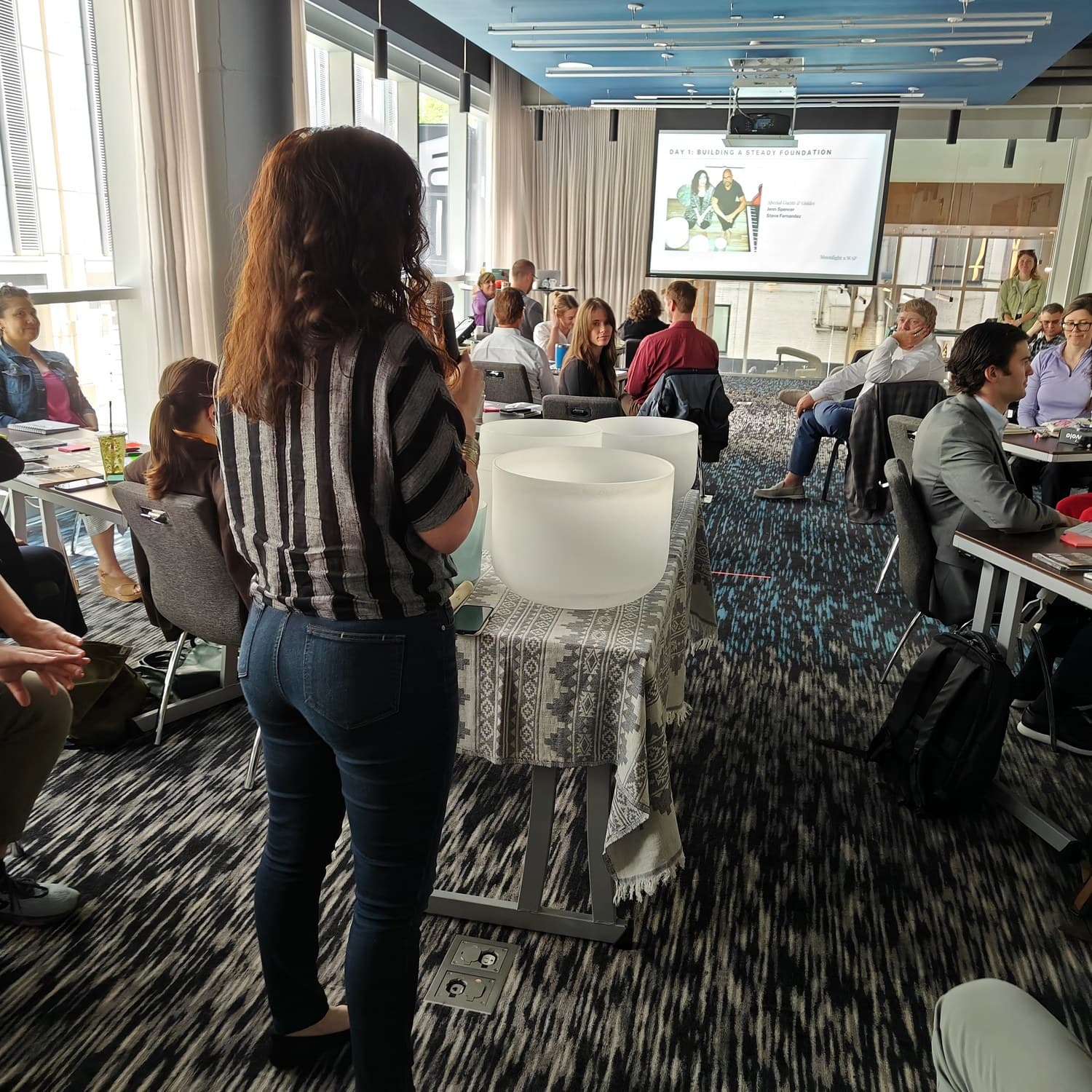 Jennifer with her back to camera, microphone in hand, facing a room of people seated at tables